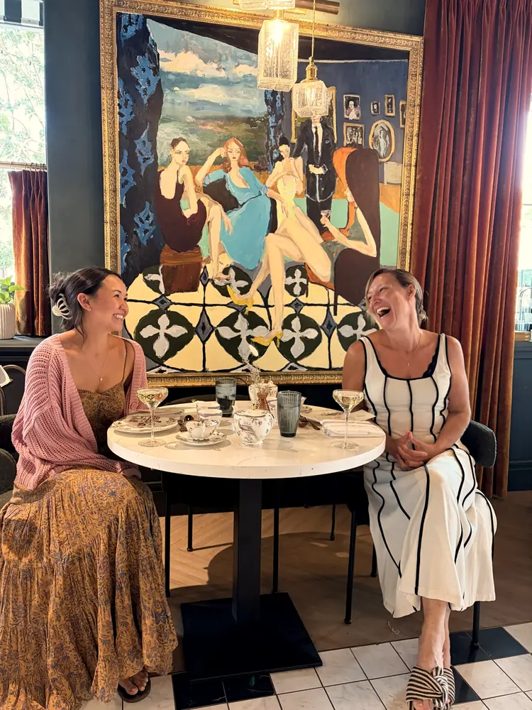 Two women laughing together over tea beneath a large painting at a gathering.