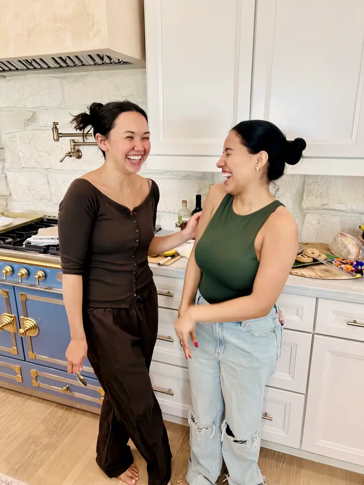Two women laughing together in the kitchen at a Becoming Beautifully gathering.